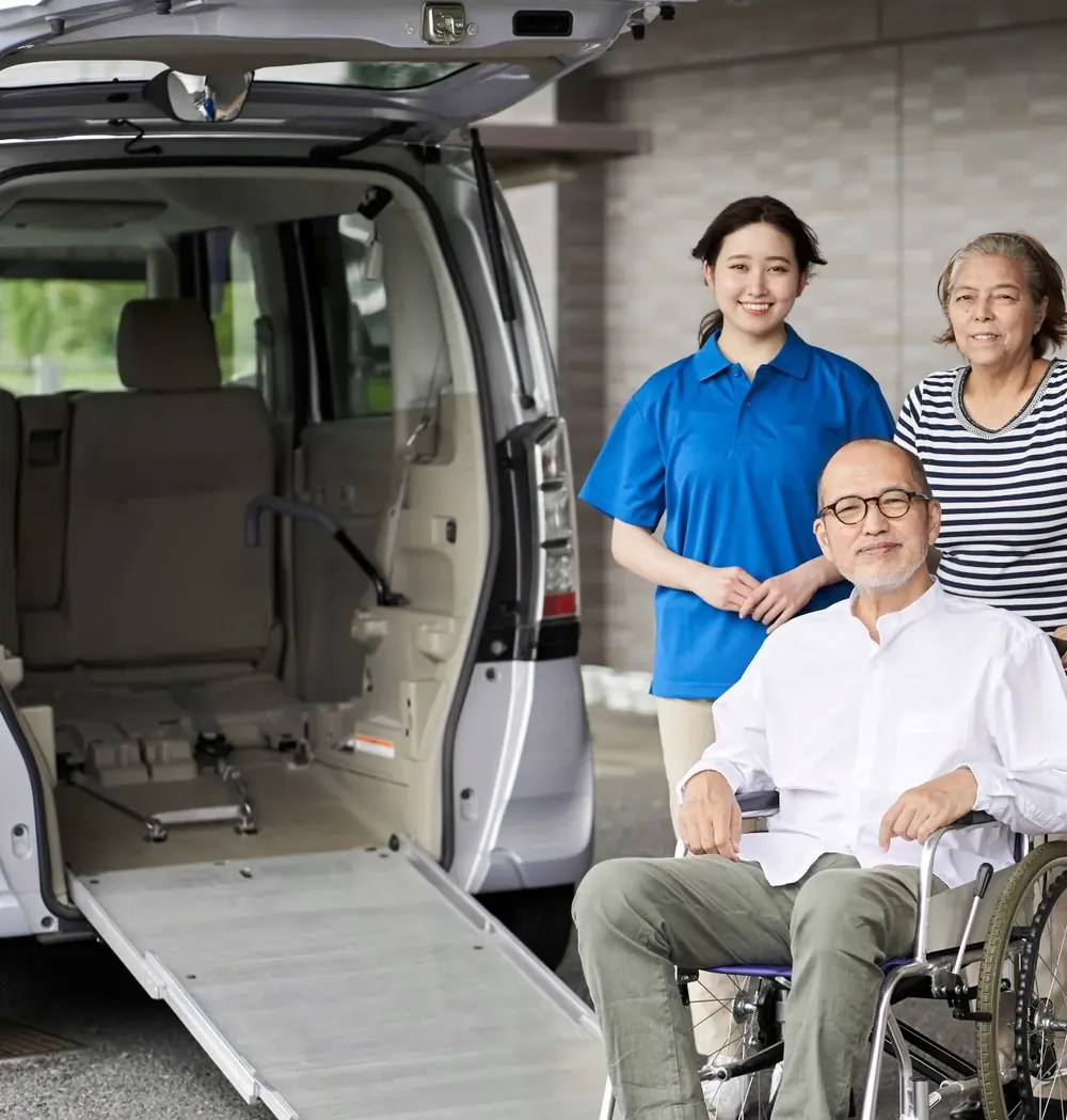 An elderly man in a wheelchair with his caregivers beside a wheelchair-accessible van